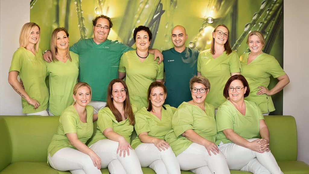 Großes, harmonisches Gruppenfoto eines medizinischen Praxisteams auf einer grünen Couch, farblich perfekt abgestimmt auf die Corporate Identity der Praxis